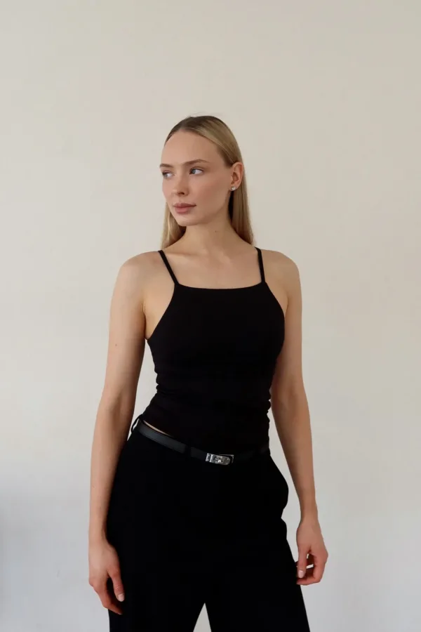 Woman in a black sleeveless dress with spaghetti straps, standing against a plain light wall, looking to her left.
