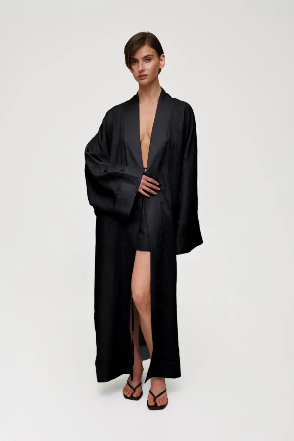 Full-body image of a woman in a black, oversized robe with a deep V opening, standing against a pale background.