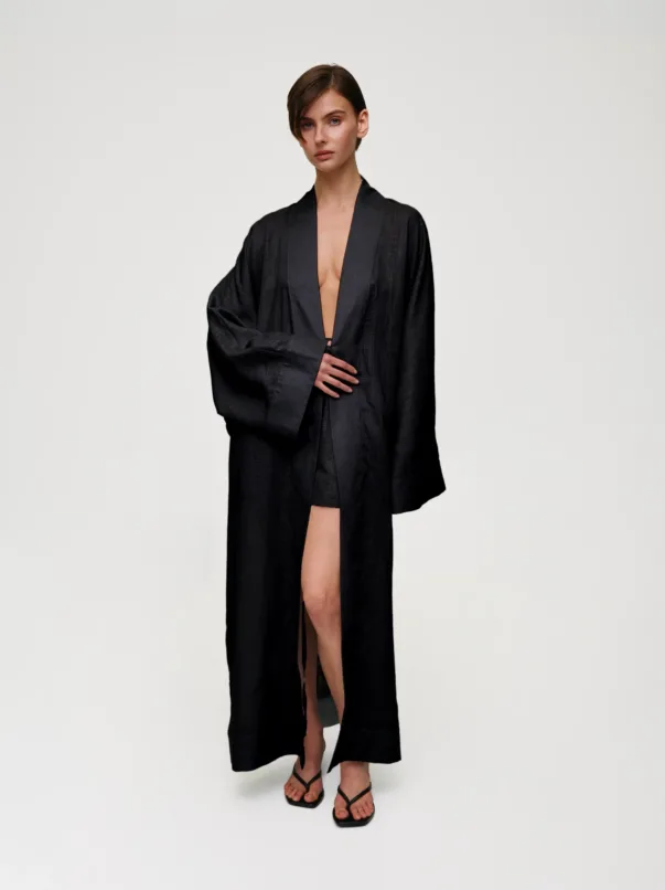 Full-body image of a woman in a black, oversized robe with a deep V opening, standing against a pale background.
