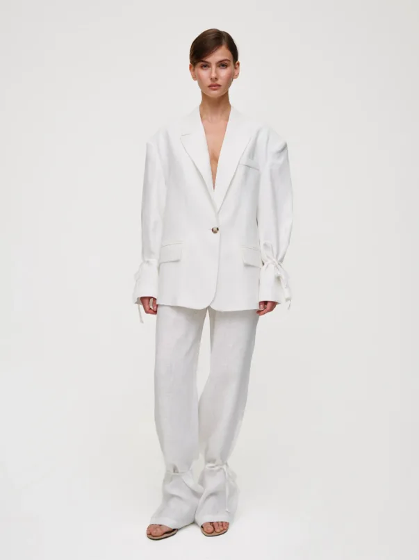 Model in an oversized white suit with wide lapels, single button, and tied cuffs, standing against a pale background.