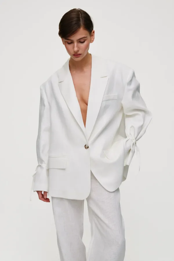 Model in a white tailored blazer and matching pants, looking downward against a plain light background.