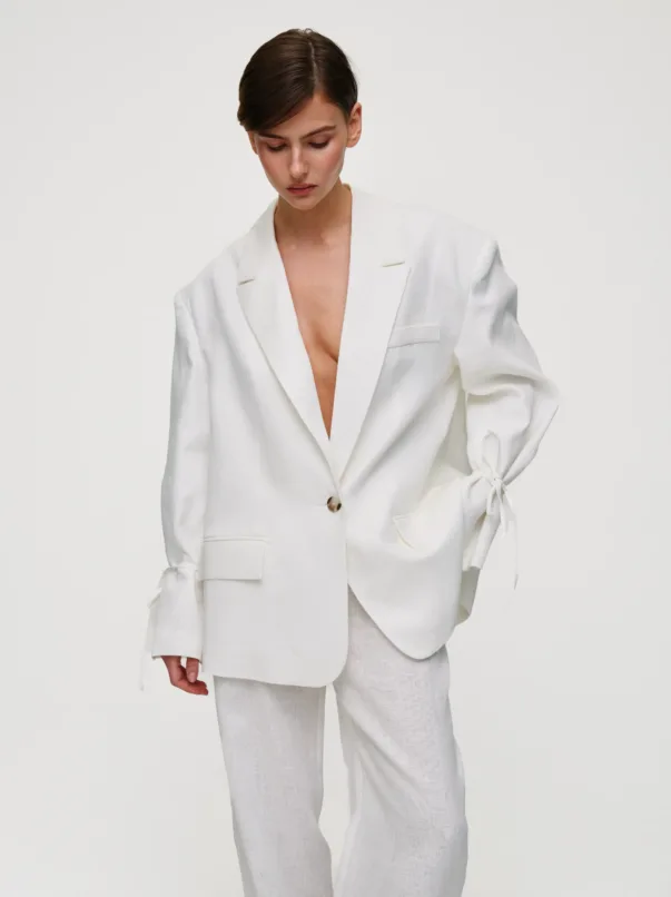 Model in a white tailored blazer and matching pants, looking downward against a plain light background.