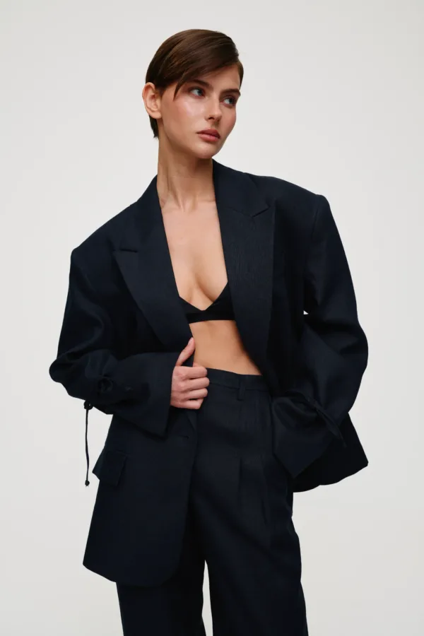 Model in a dark navy blazer and matching trousers, with a black bralette visible as she adjusts the blazer and looks to the side in a neutral studio setting.