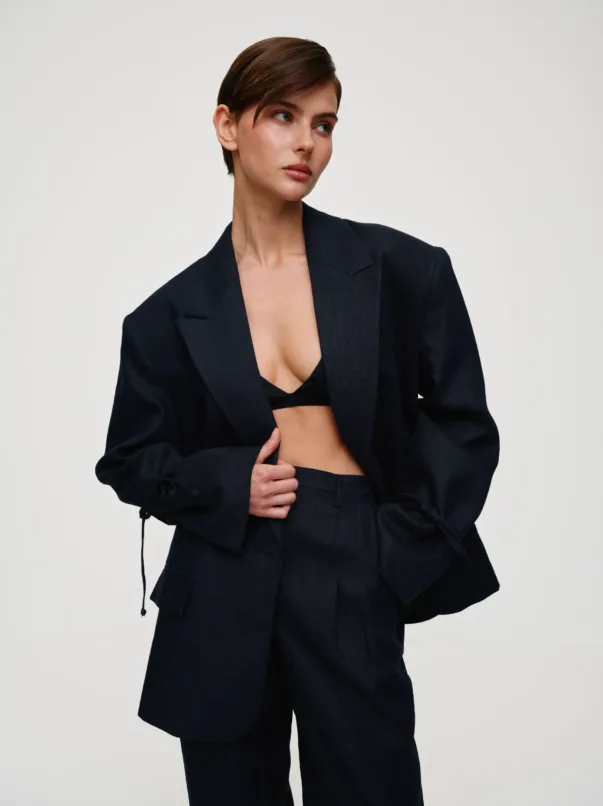 Model in a dark navy blazer and matching trousers, with a black bralette visible as she adjusts the blazer and looks to the side in a neutral studio setting.