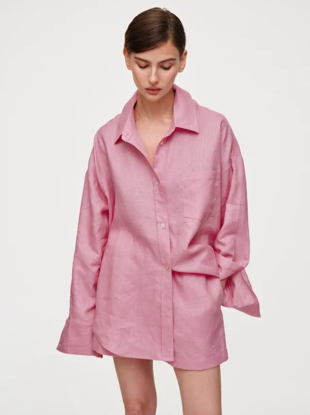 Woman in a pink oversized button-down shirt, looking downward with short dark hair against a plain light background