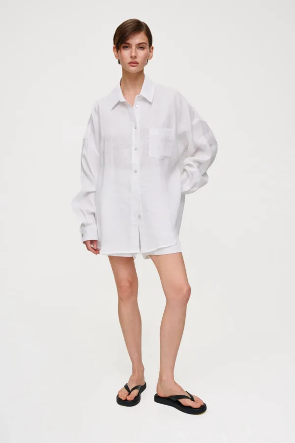 Model wearing a loose white linen shirt and matching shorts, standing against a light background in black flip-flops.