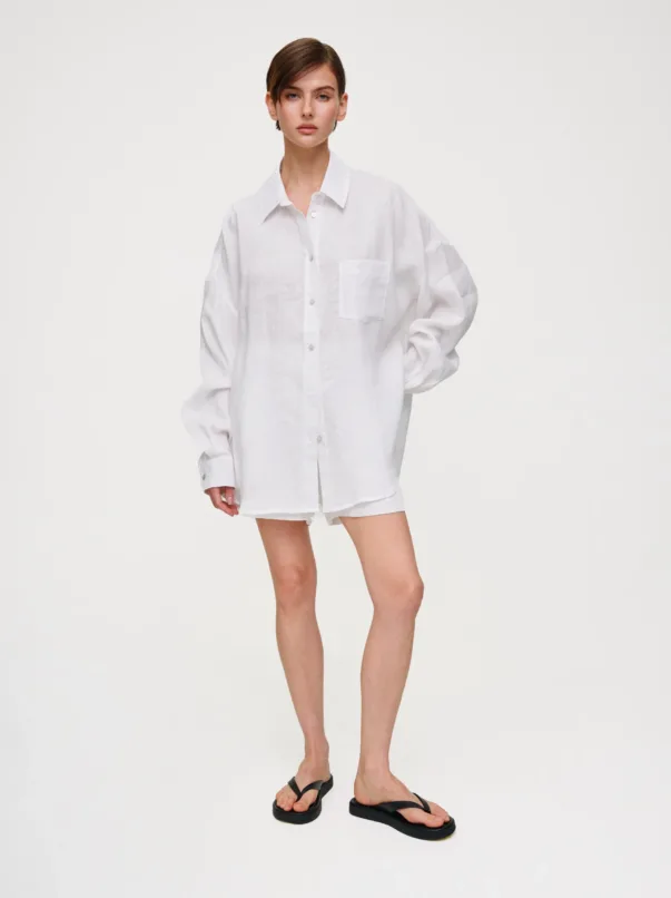 Model wearing a loose white linen shirt and matching shorts, standing against a light background in black flip-flops.