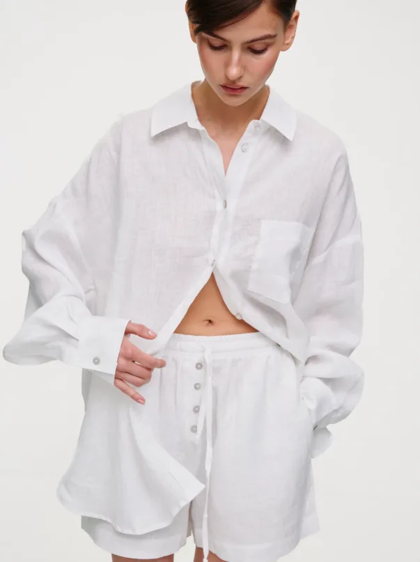 Person wearing an oversized white linen button-down shirt and matching shorts, looking down and adjusting the fabric at the waist.