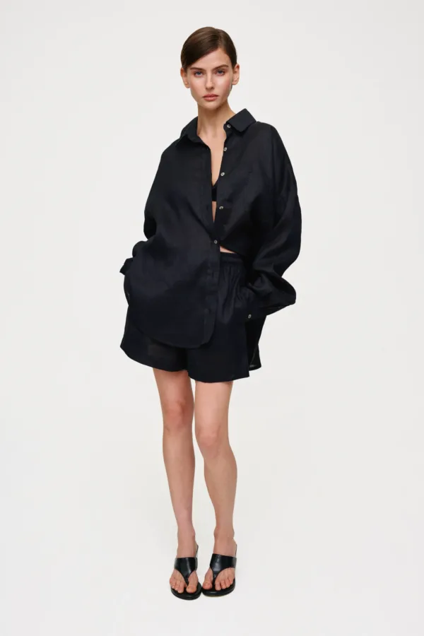 Full-body shot of a woman in an oversized black button-down shirt and matching shorts, standing against a white backdrop.