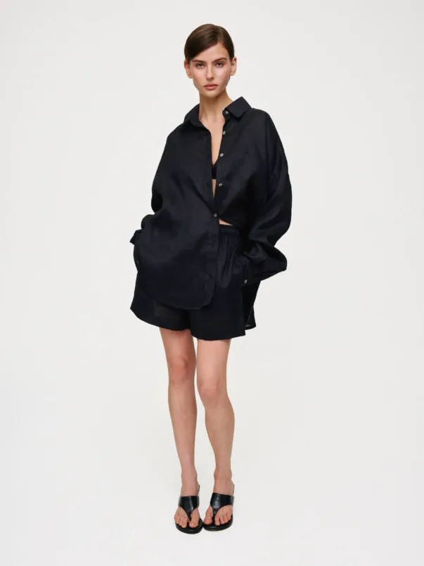 Full-body shot of a woman in an oversized black button-down shirt and matching shorts, standing against a white backdrop.