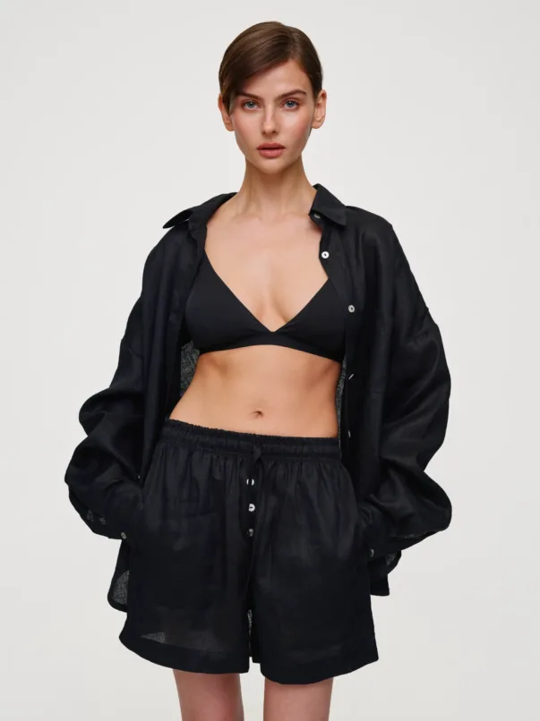 Woman wearing a black bralette with a black button-down shirt and matching shorts, posing against a light neutral background.