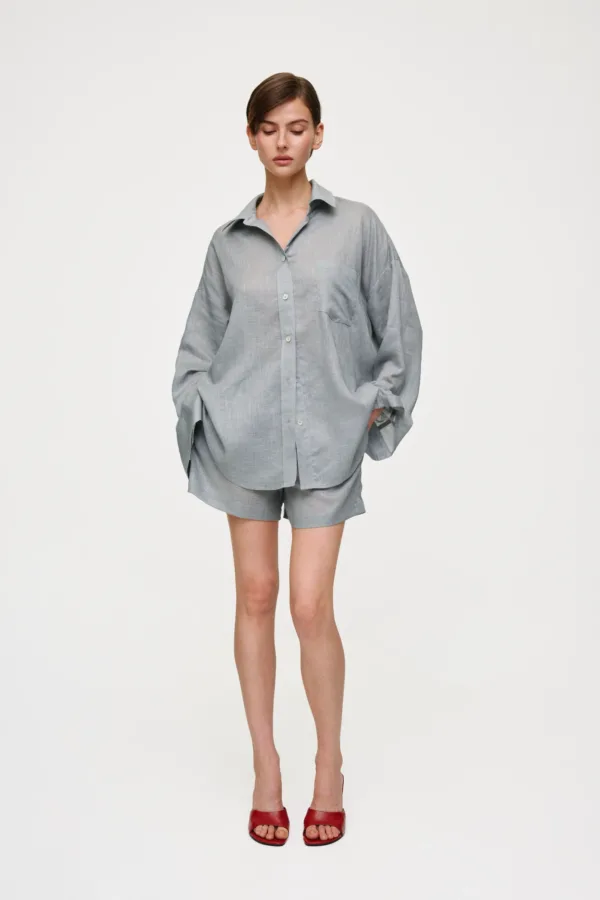 Full-body studio shot of a person in an oversized light gray shirt and matching shorts, standing with hands in pockets and wearing red sandals against a white background.