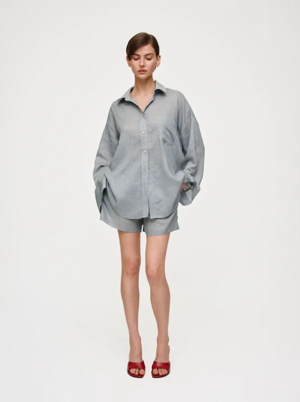 Full-body studio shot of a person in an oversized light gray shirt and matching shorts, standing with hands in pockets and wearing red sandals against a white background.