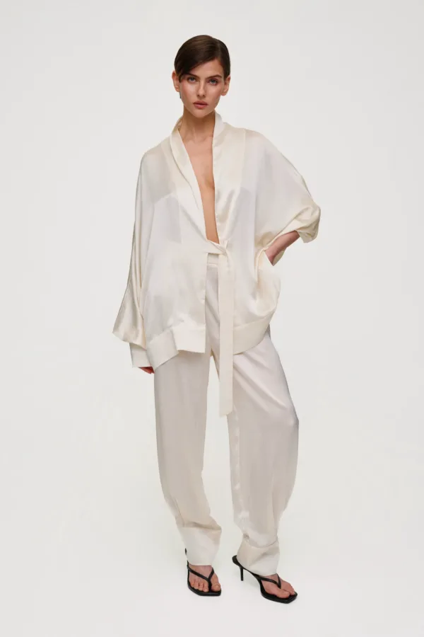 Female model wearing a cream satin robe with a belt, paired with matching wide-leg pants, and black strappy sandals against a pale background.