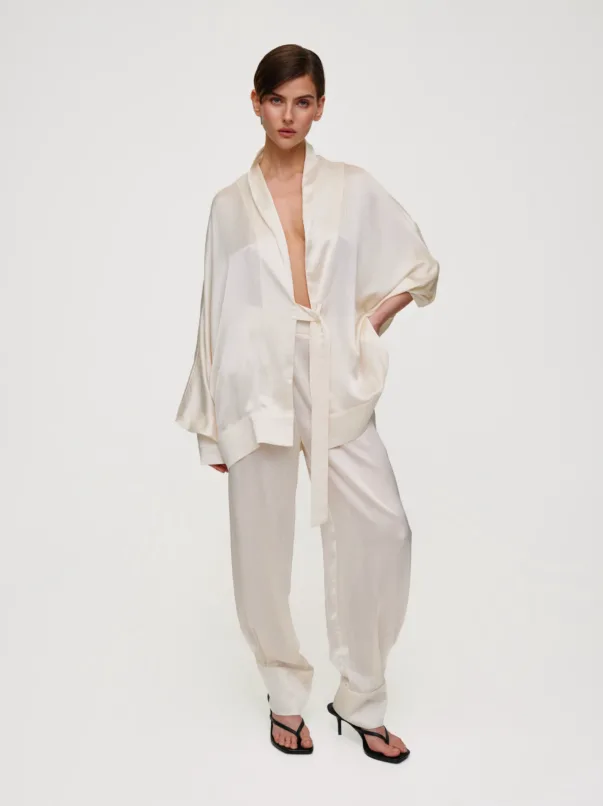 Female model wearing a cream satin robe with a belt, paired with matching wide-leg pants, and black strappy sandals against a pale background.
