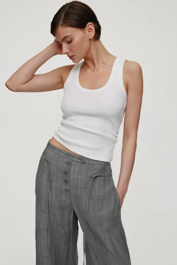 Woman with short dark hair wearing a white tank top and gray wide-leg pants, head tilted and her hand resting behind her neck.