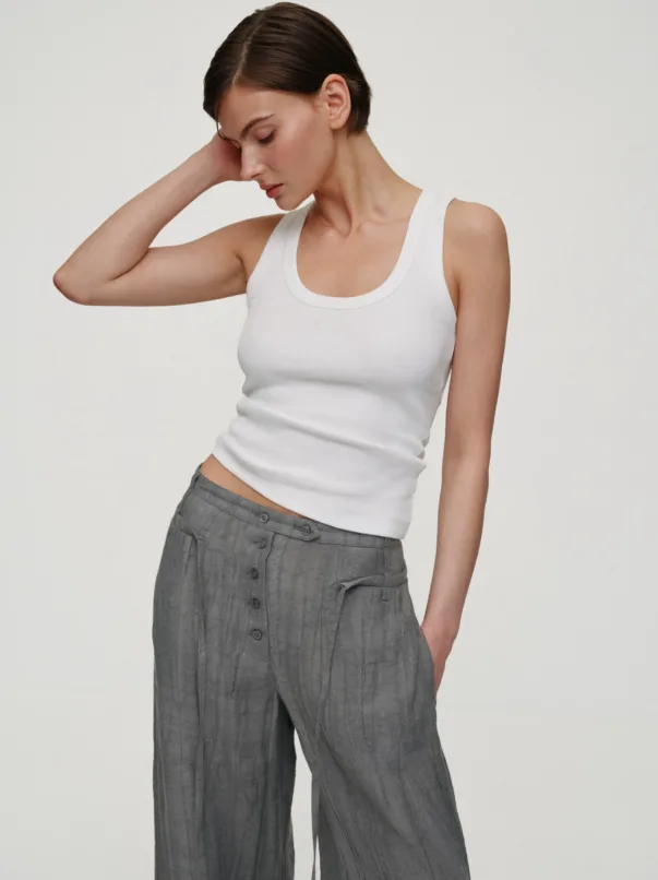 Woman with short dark hair wearing a white tank top and gray wide-leg pants, head tilted and her hand resting behind her neck.