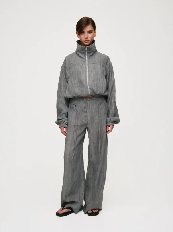 Model wearing a gray crinkled zip-front high-neck top with matching wide-leg pants, standing against a pale background.