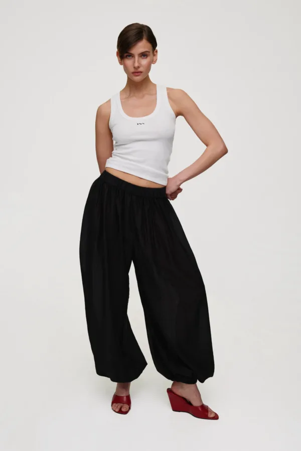 Woman posing in a white tank top and black wide-leg pants, wearing red heeled sandals, against a plain light background.