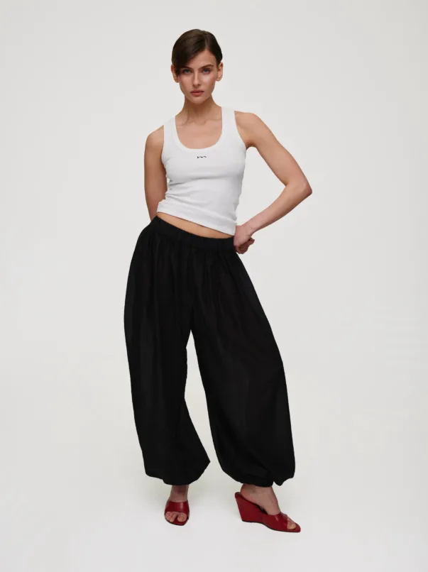 Woman posing in a white tank top and black wide-leg pants, wearing red heeled sandals, against a plain light background.
