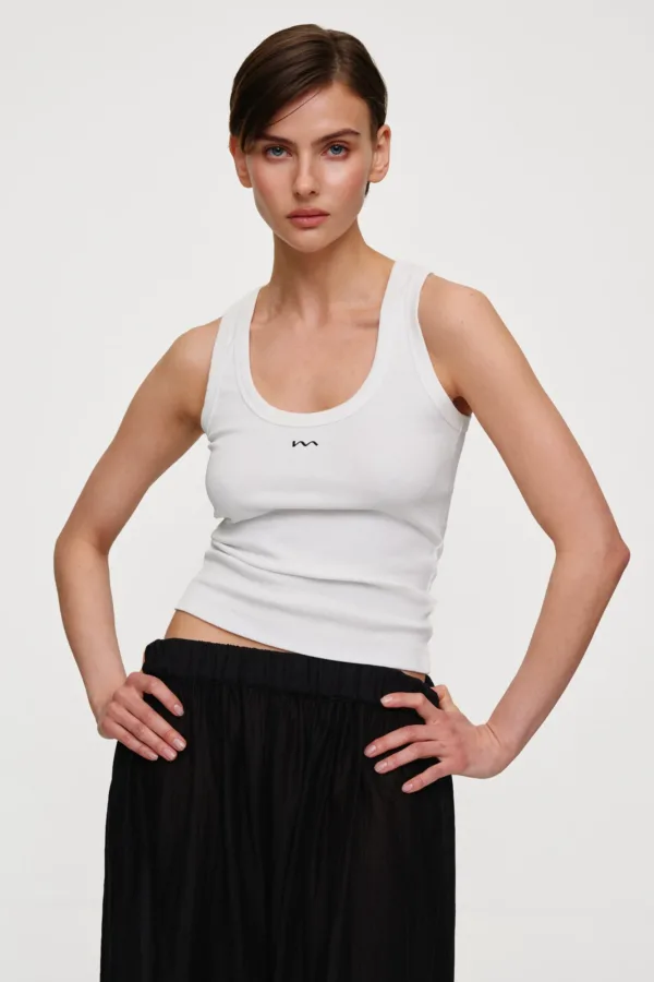 Woman posing in a white sleeveless top and black pleated skirt, hands on hips, studio backdrop.