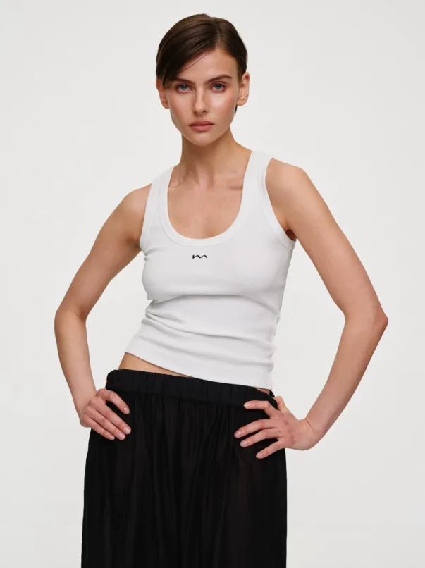 Woman posing in a white sleeveless top and black pleated skirt, hands on hips, studio backdrop.