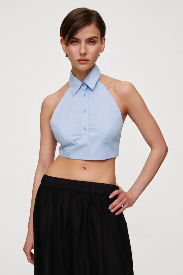 Female model wearing a light blue sleeveless halter top with a collar, paired with a black skirt, standing with one hand on her hip against a neutral background.