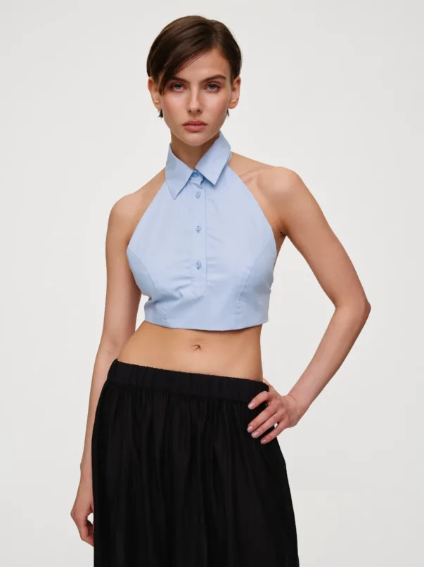 Female model wearing a light blue sleeveless halter top with a collar, paired with a black skirt, standing with one hand on her hip against a neutral background.