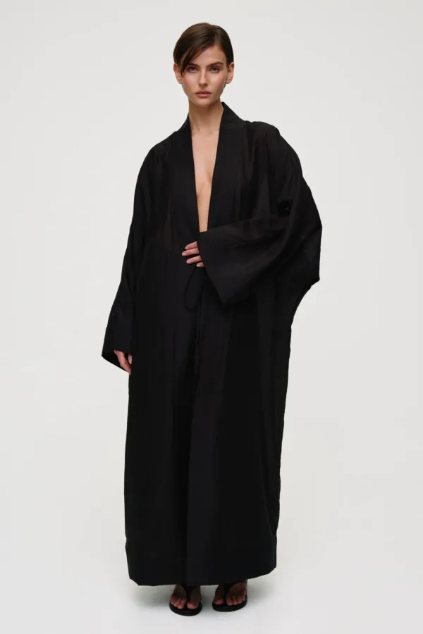 Woman modeling a long black robe with a deep neckline, standing against a light background, wearing flat black sandals.
