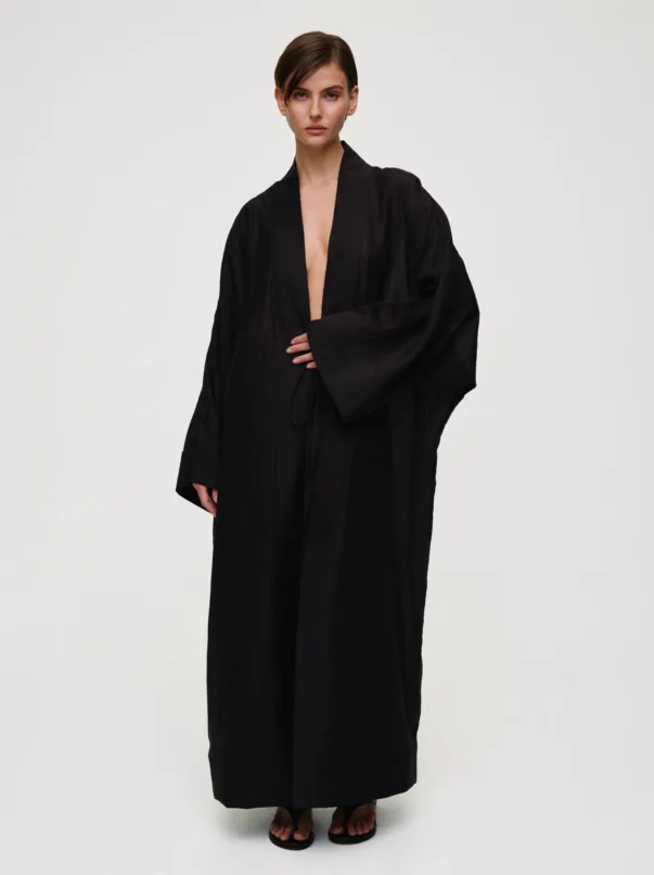Woman modeling a long black robe with a deep neckline, standing against a light background, wearing flat black sandals.