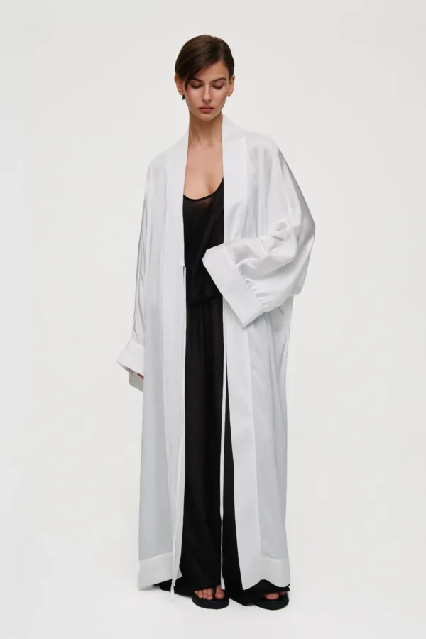 Model in a long white robe over a black outfit, standing barefoot against a pale backdrop, facing forward with a calm expression.