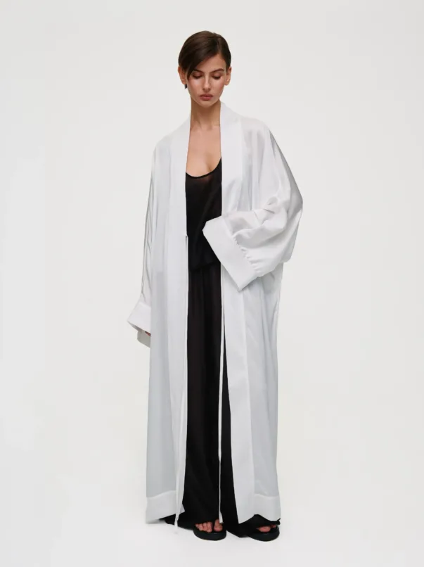 Model in a long white robe over a black outfit, standing barefoot against a pale backdrop, facing forward with a calm expression.