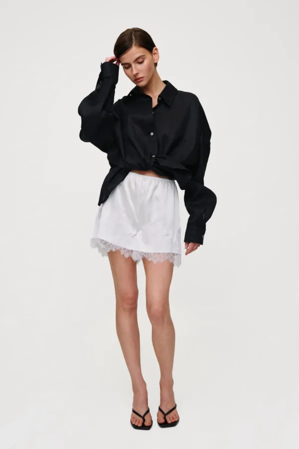 Woman in a black oversized button-down shirt knotted at the waist, a white lace mini skirt, and black sandals posing with one hand to her head.