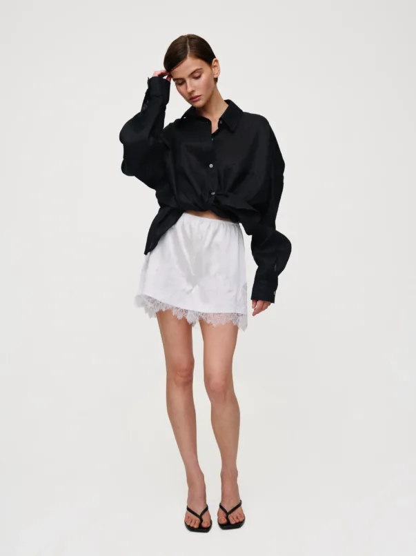Woman in a black oversized button-down shirt knotted at the waist, a white lace mini skirt, and black sandals posing with one hand to her head.