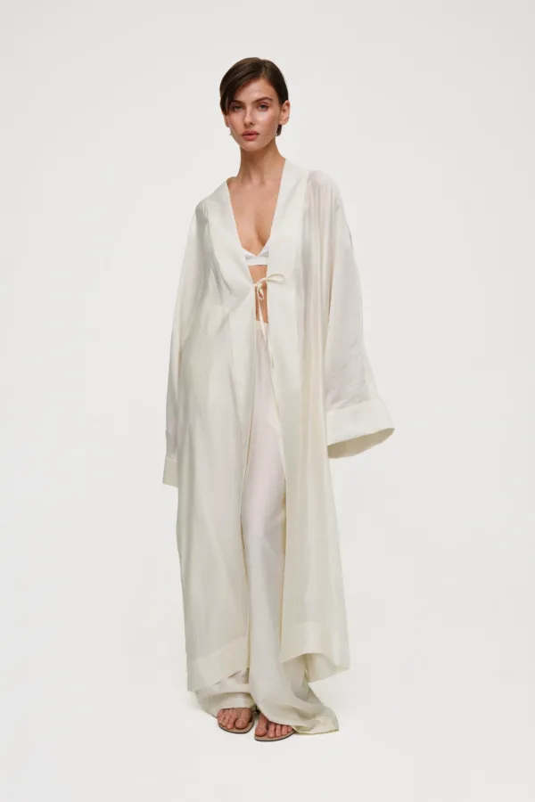 Model in a cream satin robe with a tie at the waist, standing barefoot in a studio against a white backdrop.