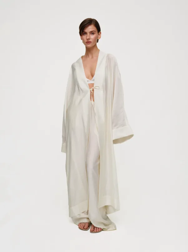 Model in a cream satin robe with a tie at the waist, standing barefoot in a studio against a white backdrop.