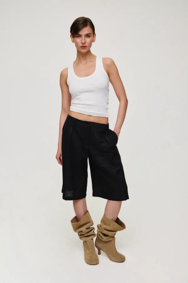 Female model wearing a white sleeveless crop top and black wide-leg shorts, paired with tan suede slouch boots, standing against a light neutral background.