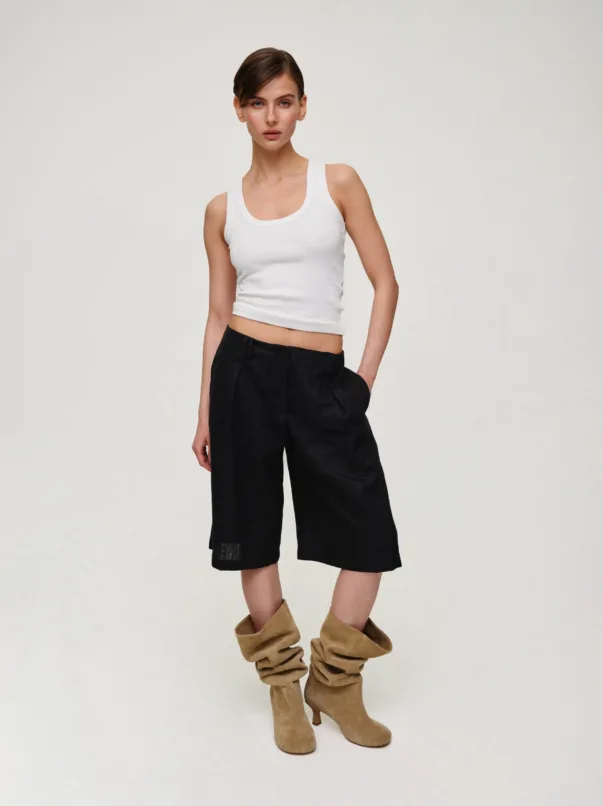 Female model wearing a white sleeveless crop top and black wide-leg shorts, paired with tan suede slouch boots, standing against a light neutral background.