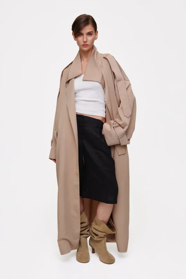 Model wearing a long beige trench over a white crop top and black skirt, with tan suede knee-high boots.