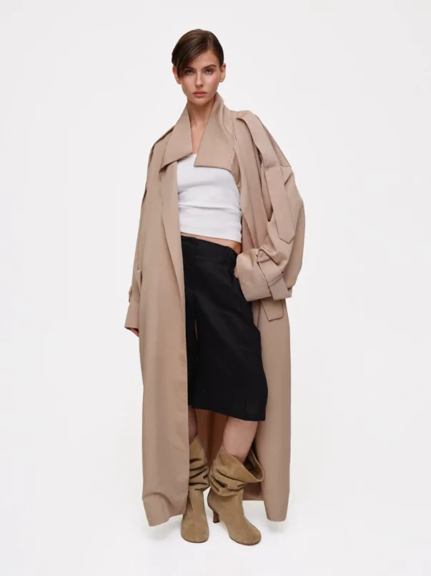 Model wearing a long beige trench over a white crop top and black skirt, with tan suede knee-high boots.
