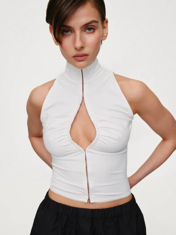 Woman wearing a white sleeveless zip-front top and black skirt, standing with hands on hips against a neutral background.