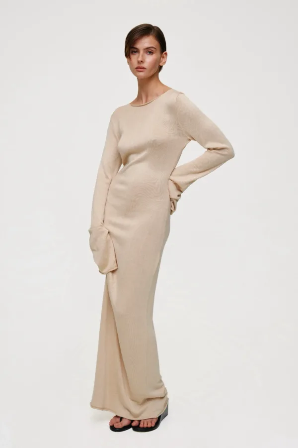 Model wearing a beige knit long-sleeve dress, standing with one hand on her hip and feet in black sandals against a light, neutral background.