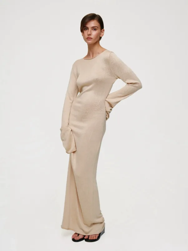 Model wearing a beige knit long-sleeve dress, standing with one hand on her hip and feet in black sandals against a light, neutral background.