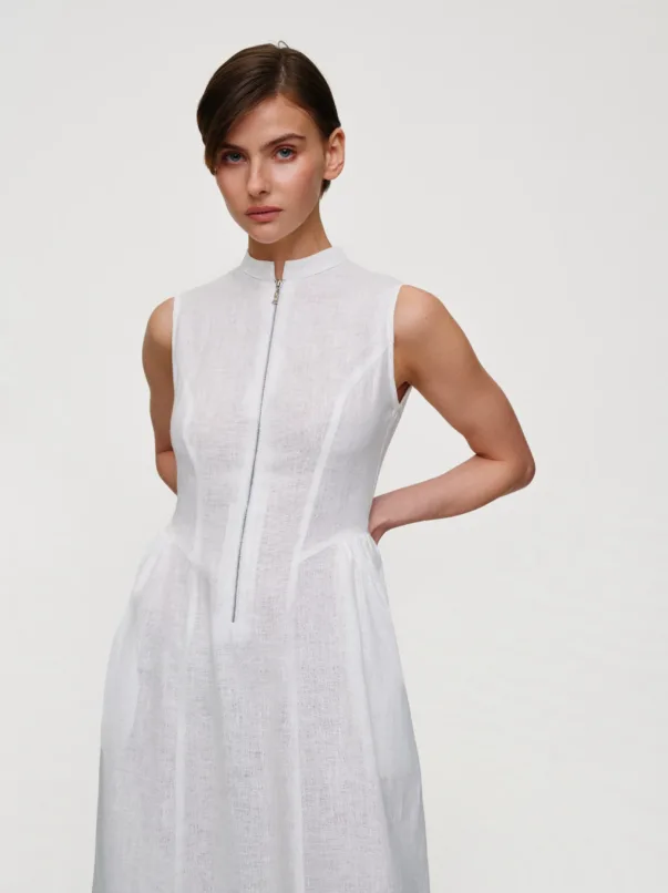 Woman in a sleeveless white dress with a front zipper, standing against a light background, looking at the camera.