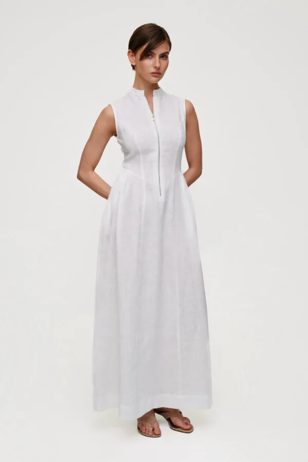 Model wearing a white sleeveless linen dress with a front zipper, standing against a pale studio background wearing tan sandals.