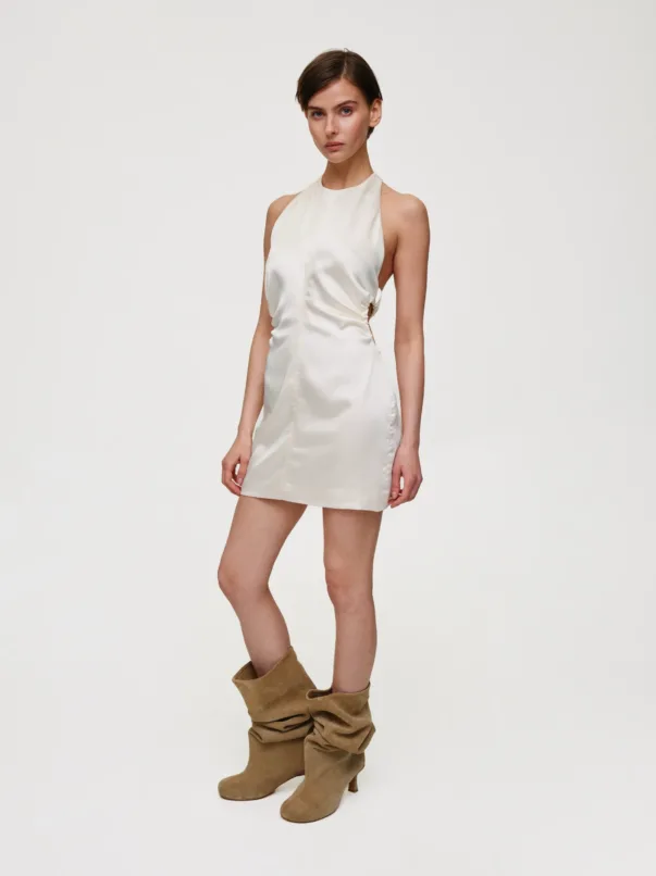 Model wearing a cream satin halter-neck mini dress and brown slouchy boots, standing against a plain light background.
