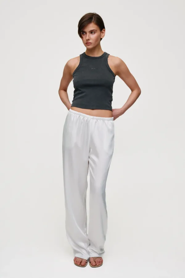Person standing against a light background, wearing a dark gray sleeveless ribbed top and white relaxed-fit pants with sandals, hands on hips.