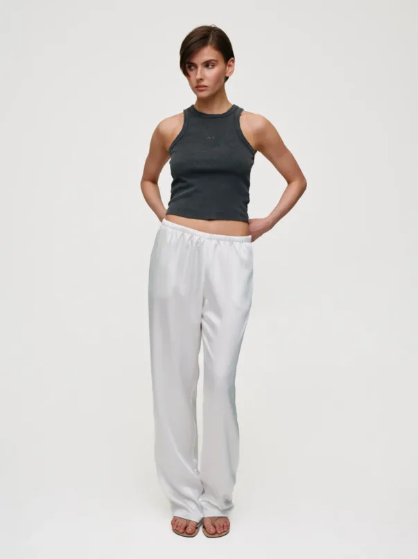 Person standing against a light background, wearing a dark gray sleeveless ribbed top and white relaxed-fit pants with sandals, hands on hips.