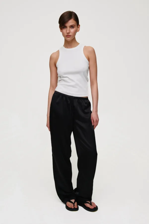 Full-body shot of a woman in a white sleeveless top and black loose-fit pants, standing with hands at her sides.
