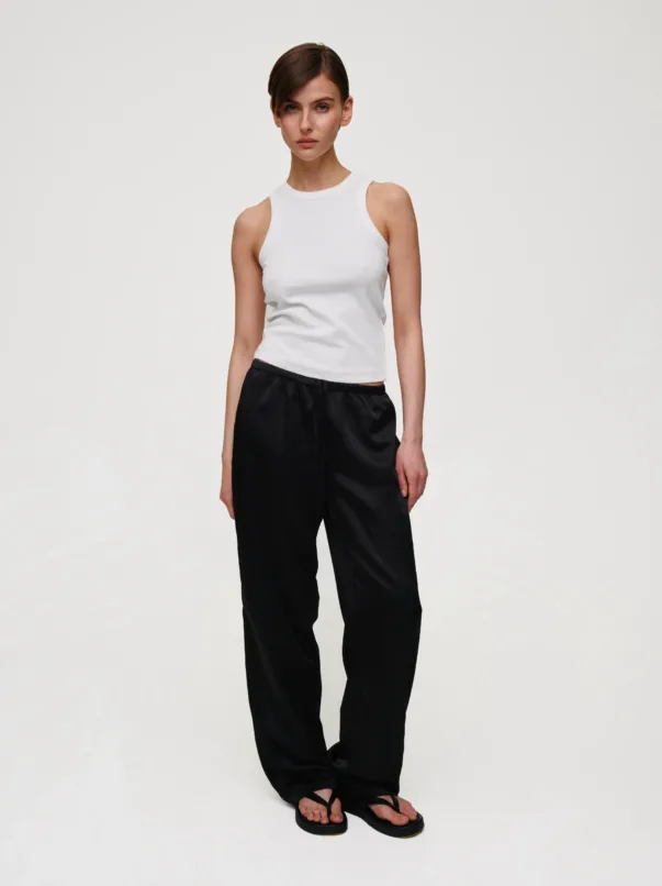Full-body shot of a woman in a white sleeveless top and black loose-fit pants, standing with hands at her sides.
